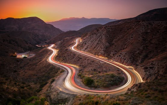 Winding mountain road with car light trails at dusk, creating scenic motion blur effect.