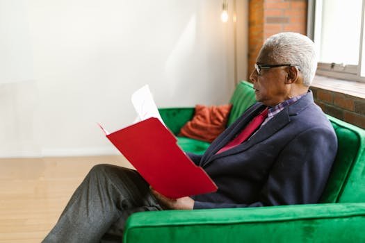 Elderly man in a blue suit reading a red folder on a vibrant green couch in a modern office setting.
