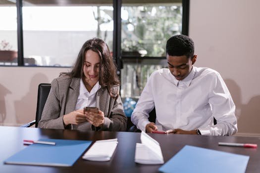 Two young professionals focused on their smartphones during an office meeting.