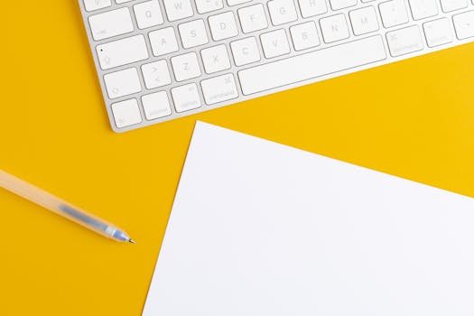 A minimalist office setup with a keyboard, pen, and paper on a vibrant yellow background.