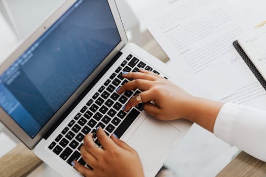 Close-up of hands typing on a laptop surrounded by papers, showcasing modern work setup.