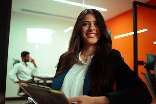 Smiling young businesswoman holding a tablet in a brightly lit office, orange wall in background.