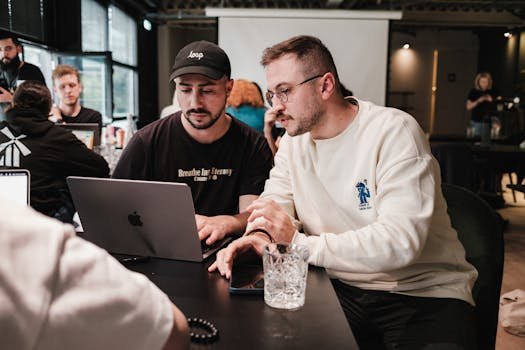 Two men collaborating on a project in a modern caf&eacute; setting with laptops and glasses.