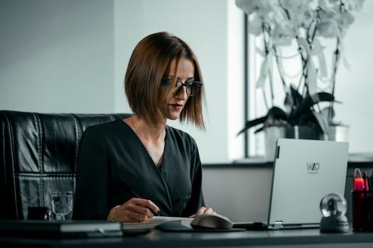 Businesswoman in an office setting, focused on work using a laptop. Elegant and professional atmosphere.