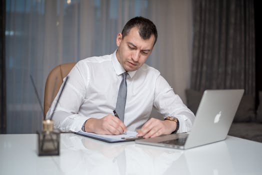 Businessman in suit writing notes at desk with laptop, reflecting modern office life.