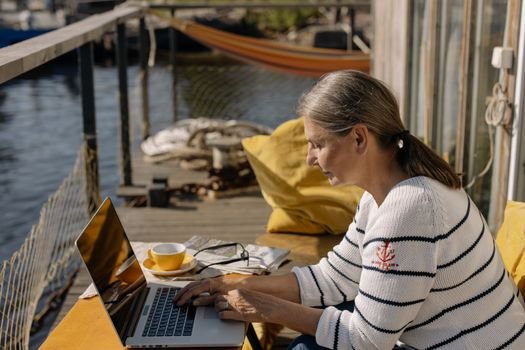 Elderly woman using a laptop on a sunny patio by the water, showcasing remote work