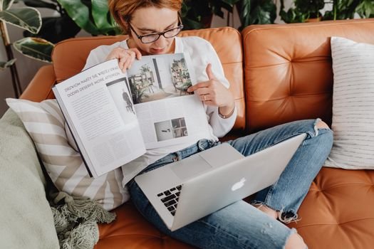 Woman reading a magazine on a brown leather sofa while using a laptop.