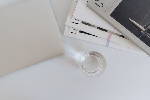 A clean and minimalist workspace featuring a laptop, magazines, and a glass on a white desk.