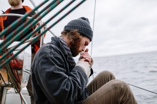 A man sitting and lighting a cigarette on a sailboat with the ocean in the background.