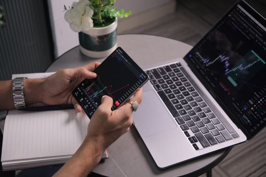 Man analyzing stock trends with laptop and mobile trading app at a modern workspace.