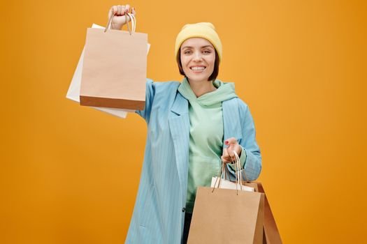 A cheerful woman in a beanie and blue coat holding shopping bags, against a vibrant yellow background.